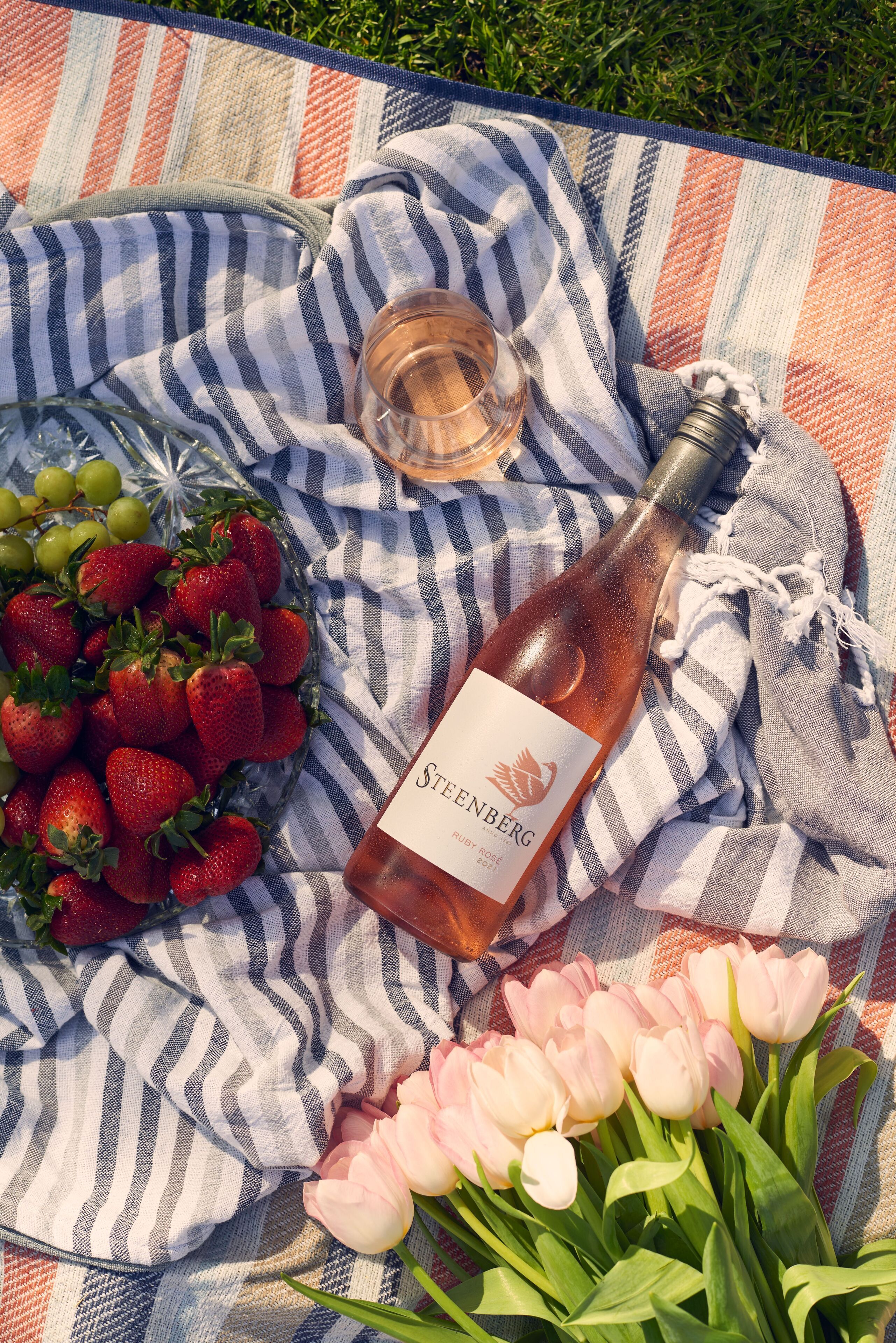 Un montaje para picnic con una botella de vino rosado, fresas y tulipanes sobre un mantel a rayas.