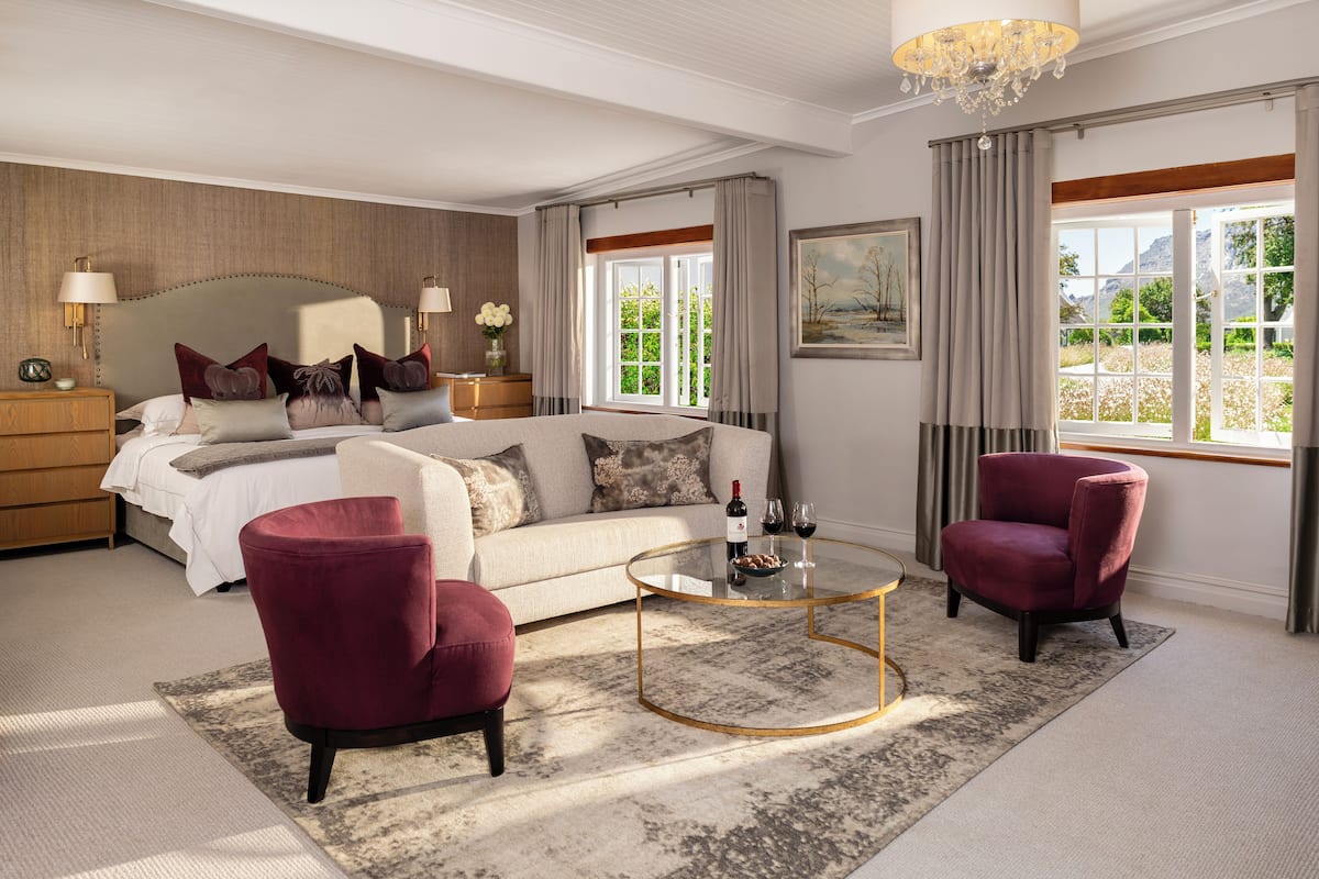 A spacious bedroom with a large bed, two armchairs, a round coffee table, and a view of greenery through the windows.