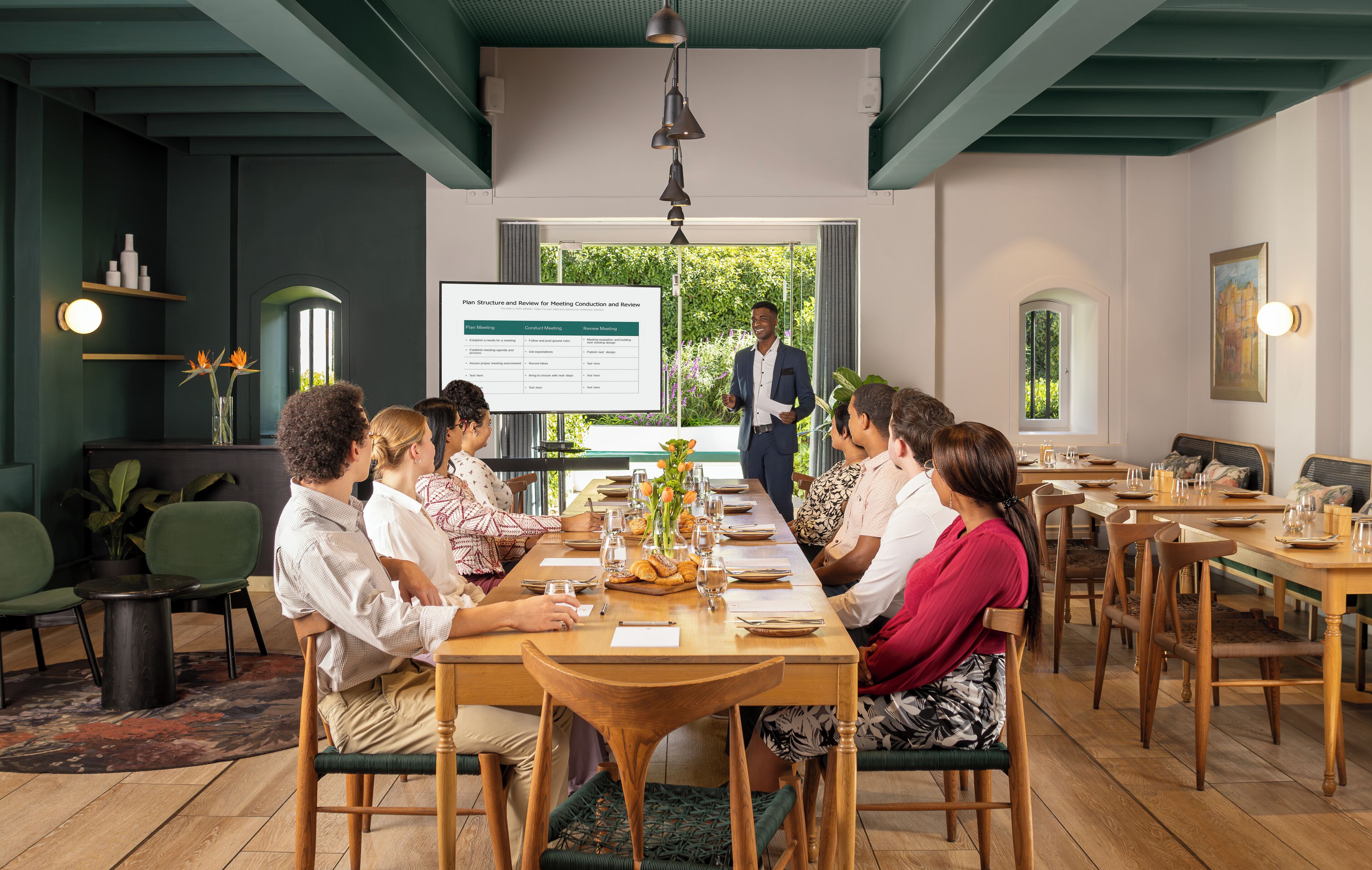 A meeting room with a long table, chairs, a projector screen, and a person presenting.