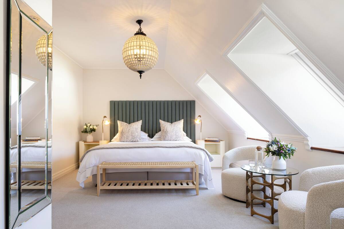 A bedroom with a bed, bedside tables, a bench, a chair, a glass table, and a chandelier.