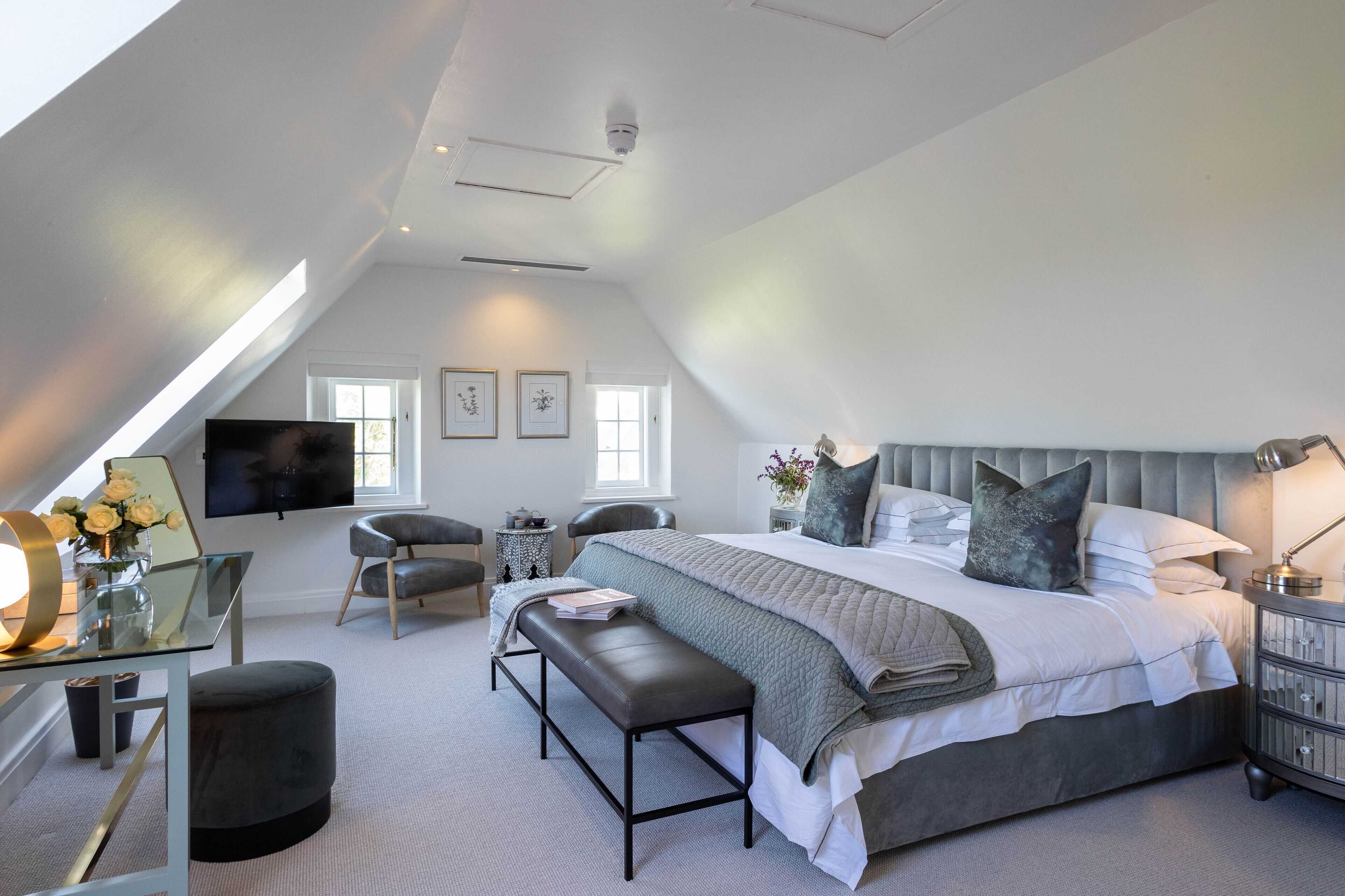 A modern bedroom with a large bed, a desk with a mirror, a bench, and a TV mounted on the wall.