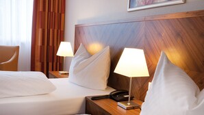 Habitación de hotel con dos camas, cada una con ropa de cama y almohadas blancas, cabeceros de madera, lámparas de noche, teléfono y un cuadro en la pared.