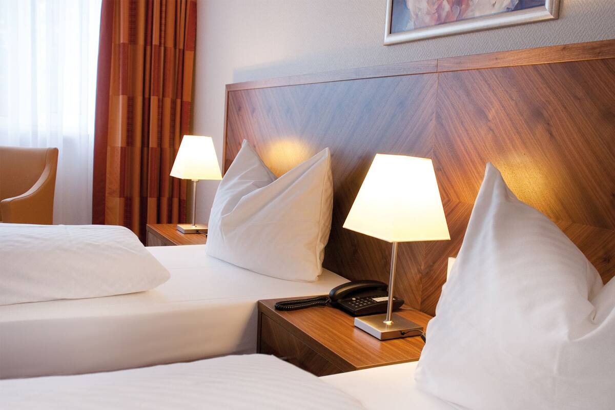 A hotel room with two beds, each with white linens and pillows, a wooden headboard, bedside lamps, a telephone, and a framed picture on the wall.