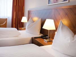 Habitación de hotel con dos camas, cada una con ropa de cama y almohadas blancas, cabeceros de madera, lámparas de noche, teléfono y un cuadro en la pared.