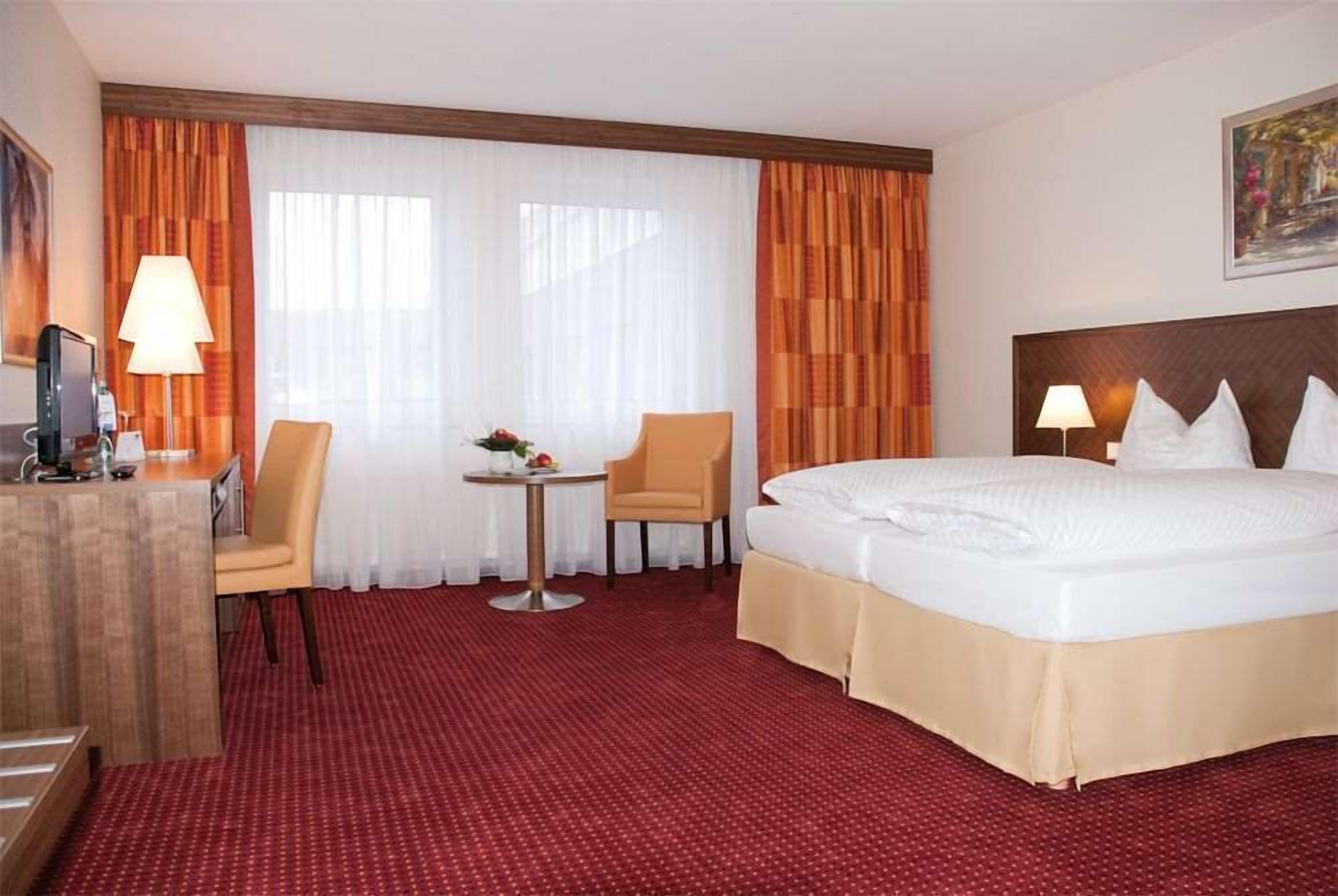 A hotel room with a large bed, two chairs, a small table with a lamp, and a desk with a lamp.
