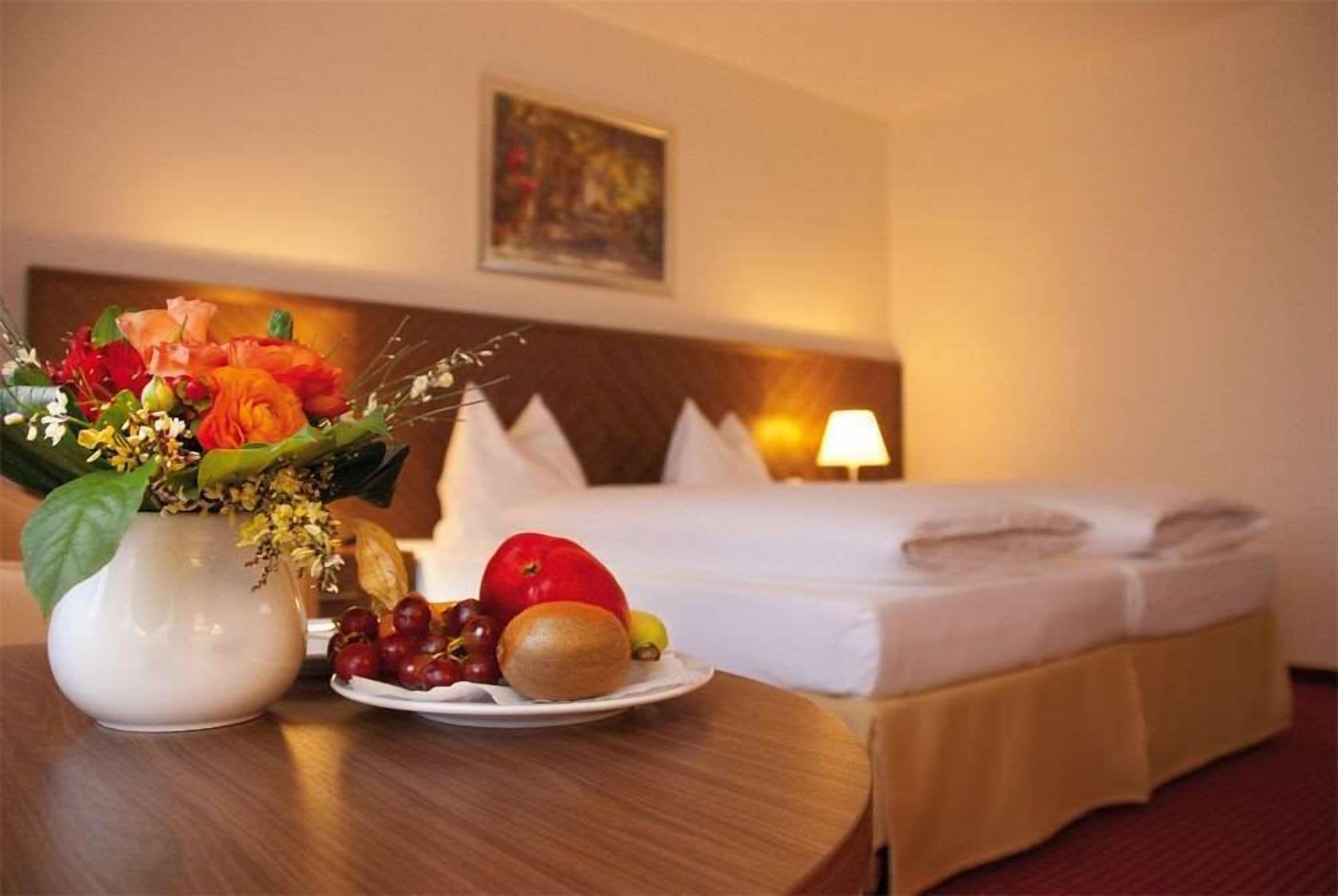 Een hotelkamer met een net opgemaakt bed, een bedlampje, een vaas met bloemen en een schaal fruit.