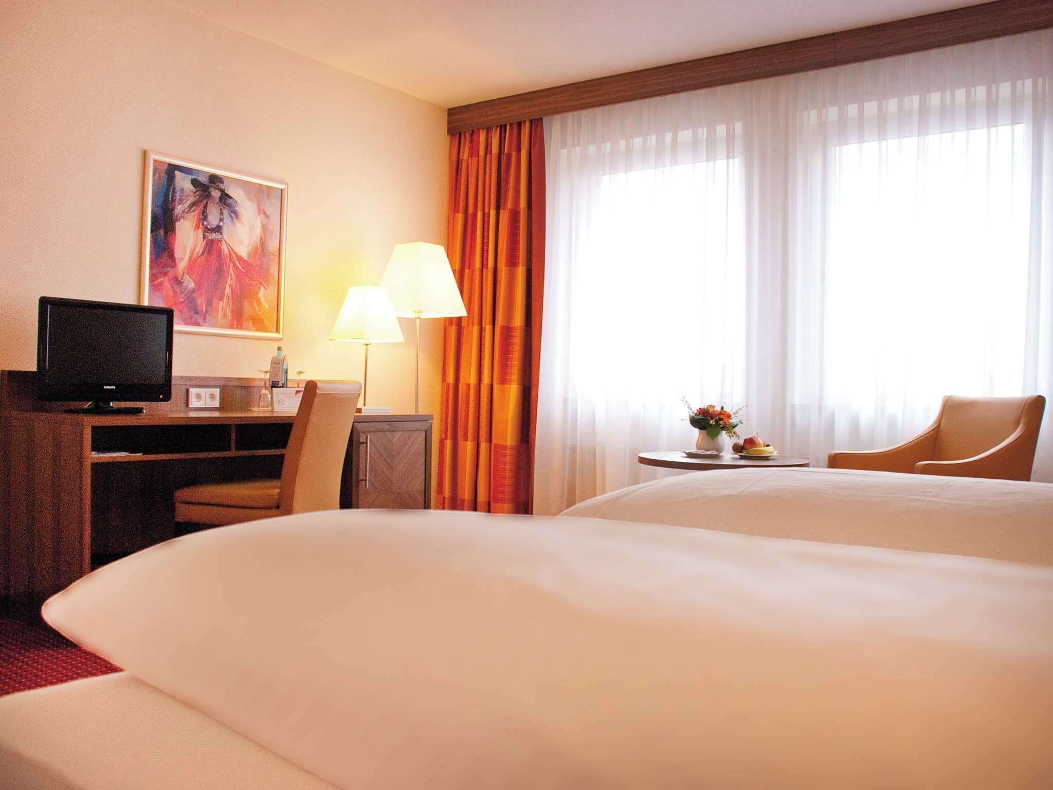A hotel room with two beds, a desk with a computer, a chair, a lamp, and a painting on the wall.