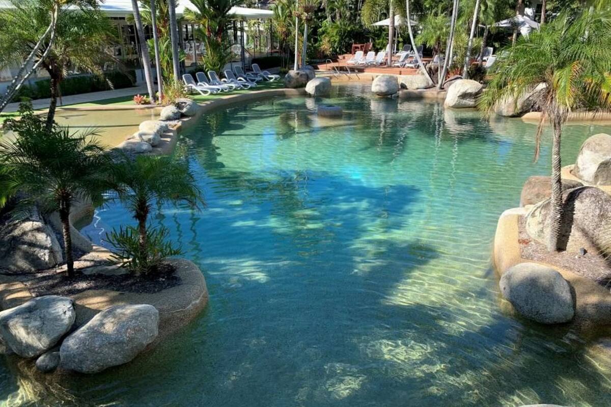 A clear pool with rocks and palm trees, surrounded by lounge chairs and umbrellas.