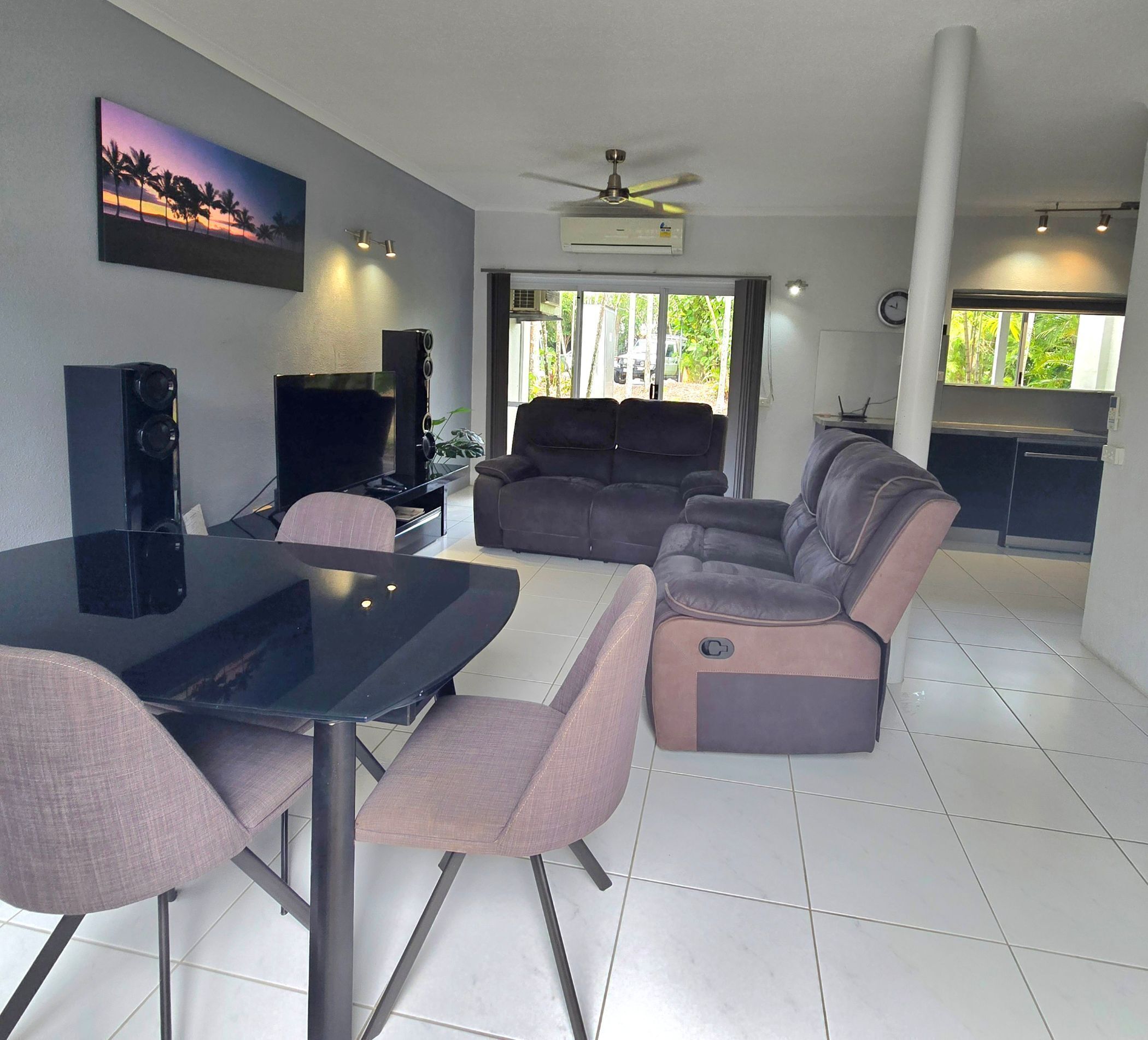 A modern living room with a flat-screen TV, a sofa, a dining table with chairs, and a ceiling fan.