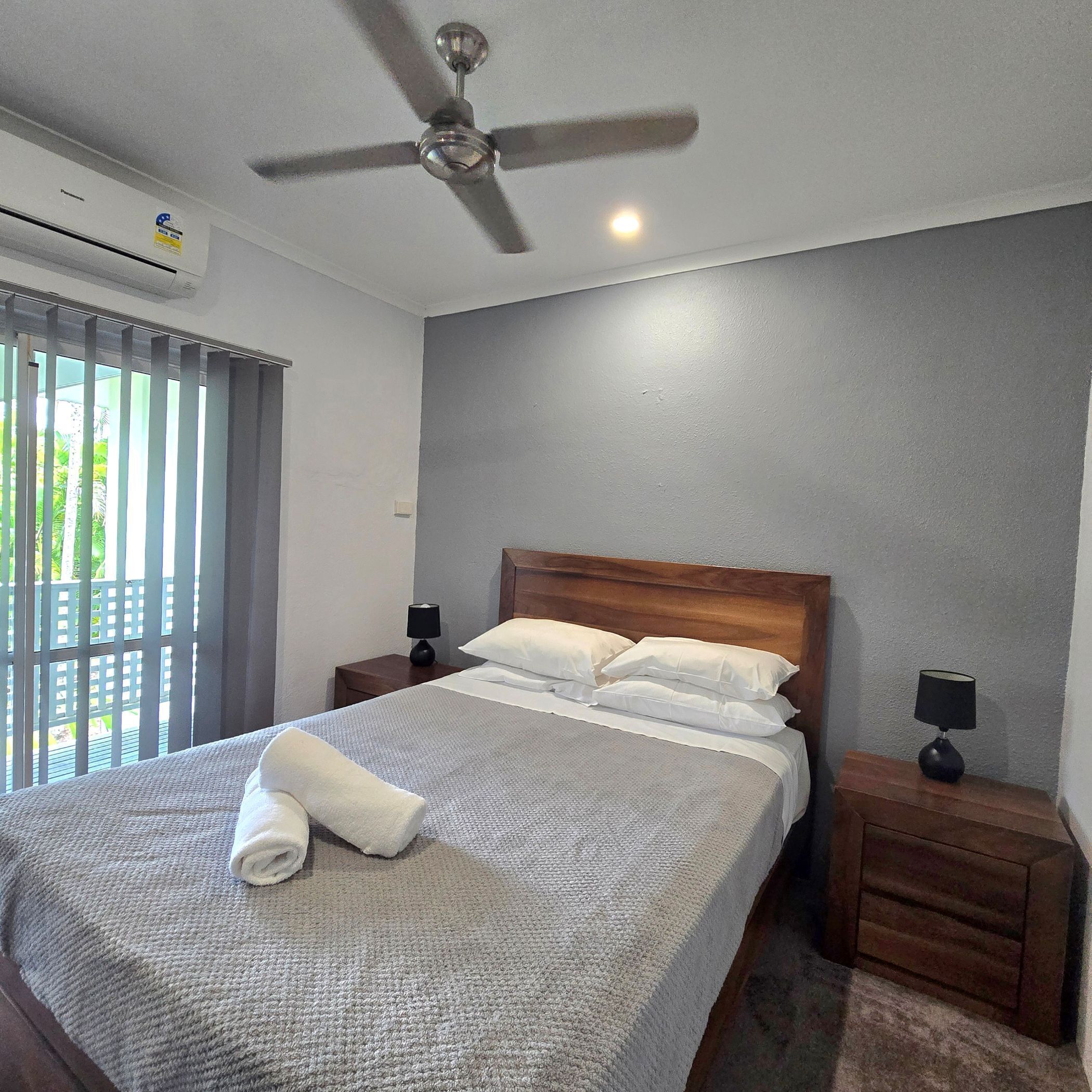 Quarto de hotel com cama, ventilador de teto, mesa de cabeceira com luminária e janela com persianas.