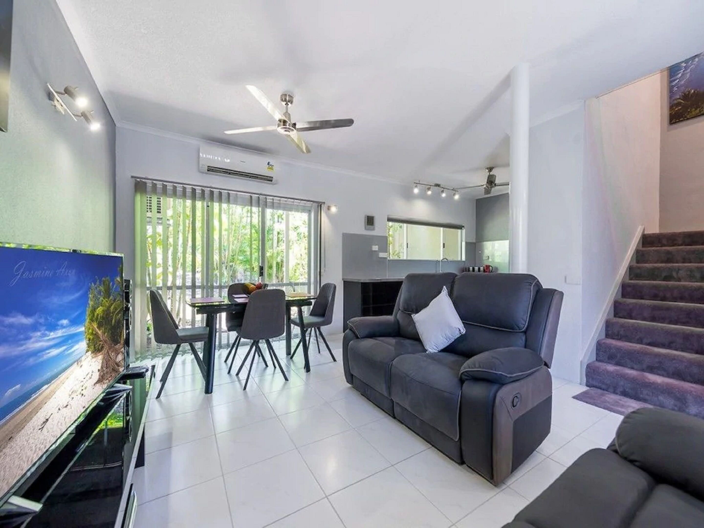 A modern living room with a sofa, dining table, and chairs. There is a TV on the left and a staircase on the right.