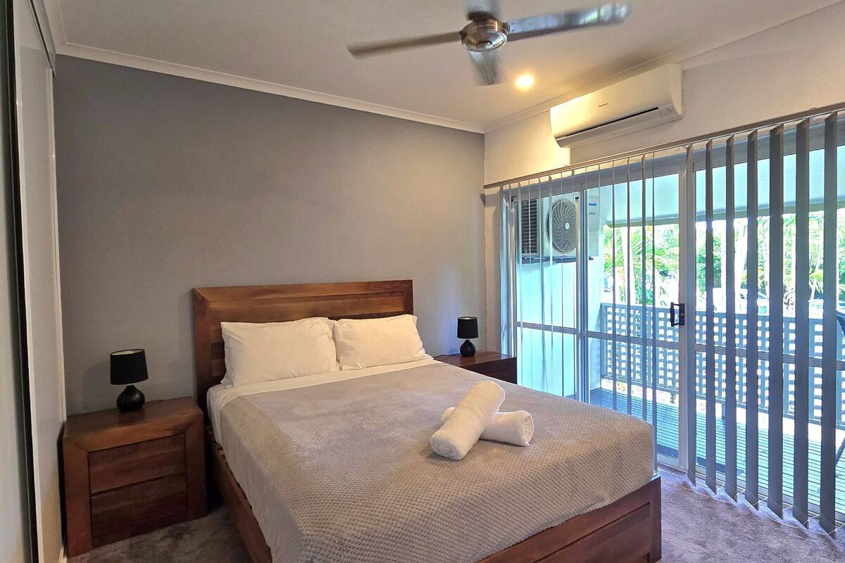 A bedroom with a bed, two bedside tables, a ceiling fan, an air conditioning unit, and a view of the outdoors through a window with a metal gate.