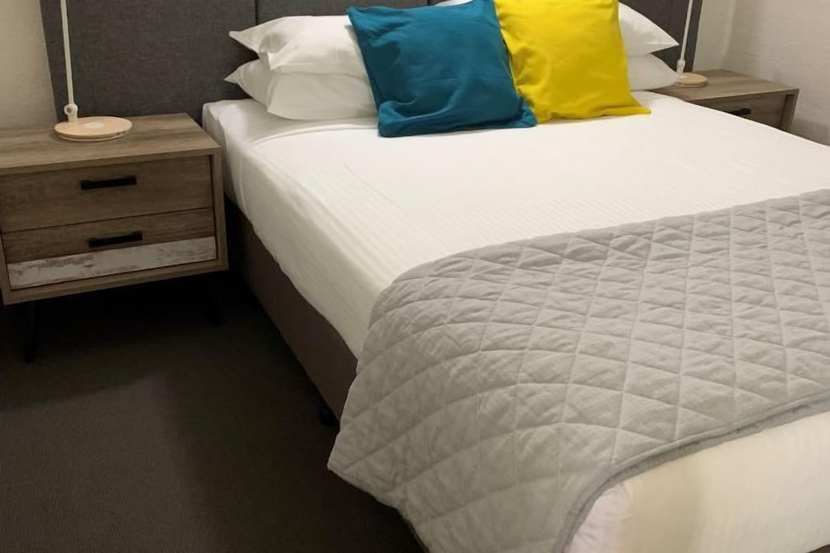 Een netjes opgemaakt bed met een witte dekbedovertrek en een grijze gewatteerde deken. Er zijn twee nachtkastjes, waarvan één met een lade en de ander met een lamp.