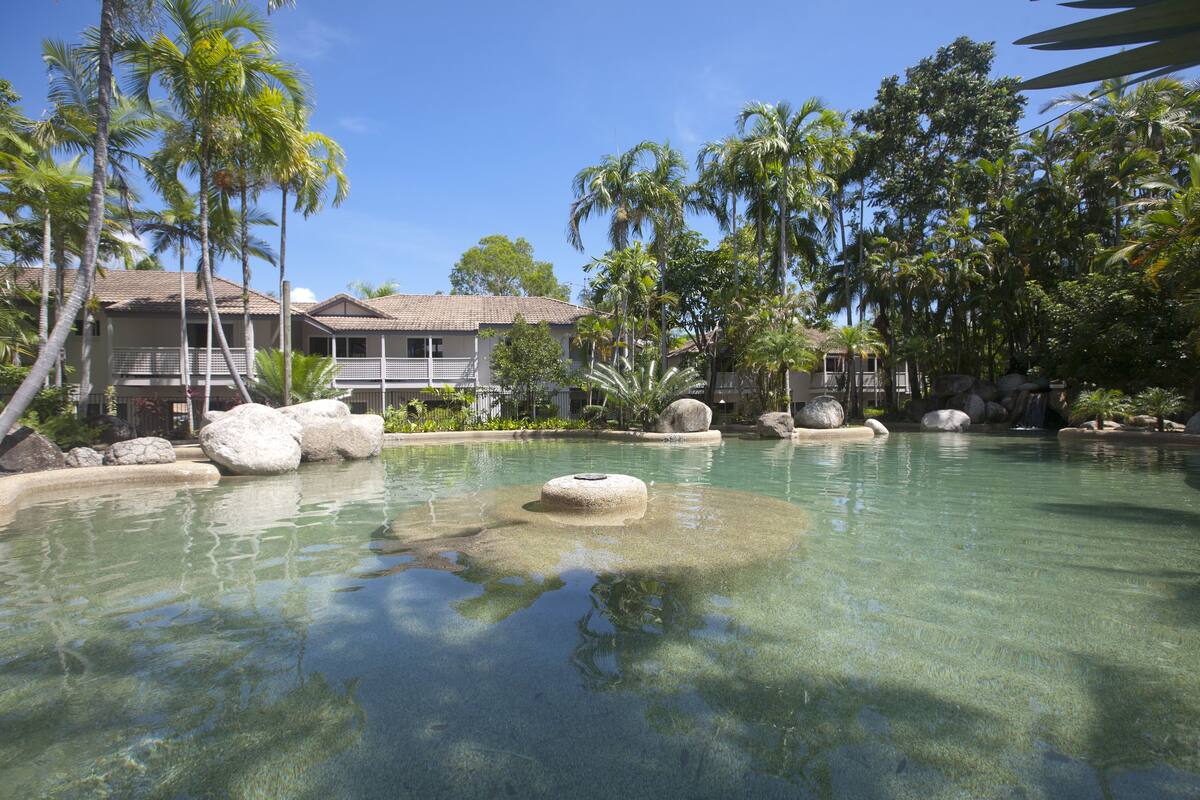 A clear pool with rocks, surrounded by palm trees and a building with a tiled roof.