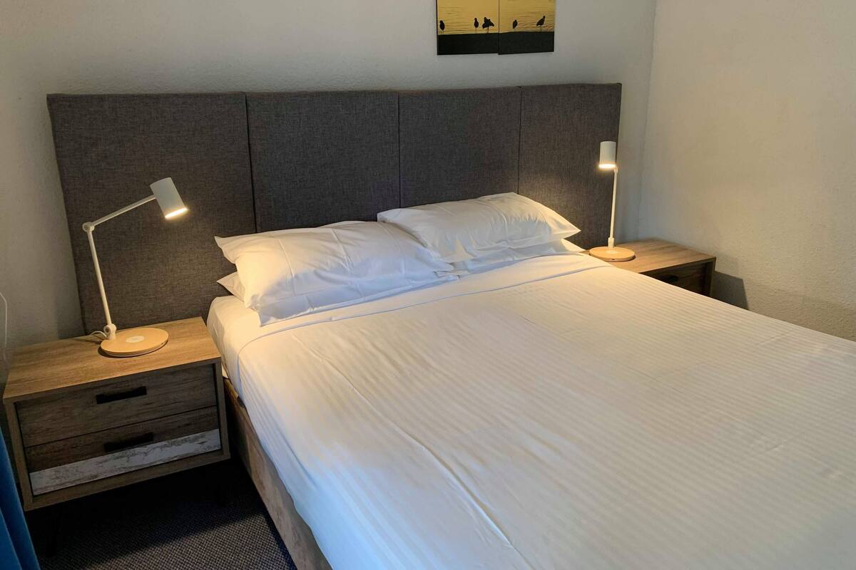 A neatly made bed with white linens, two bedside tables with lamps, and a painting of birds on the wall.