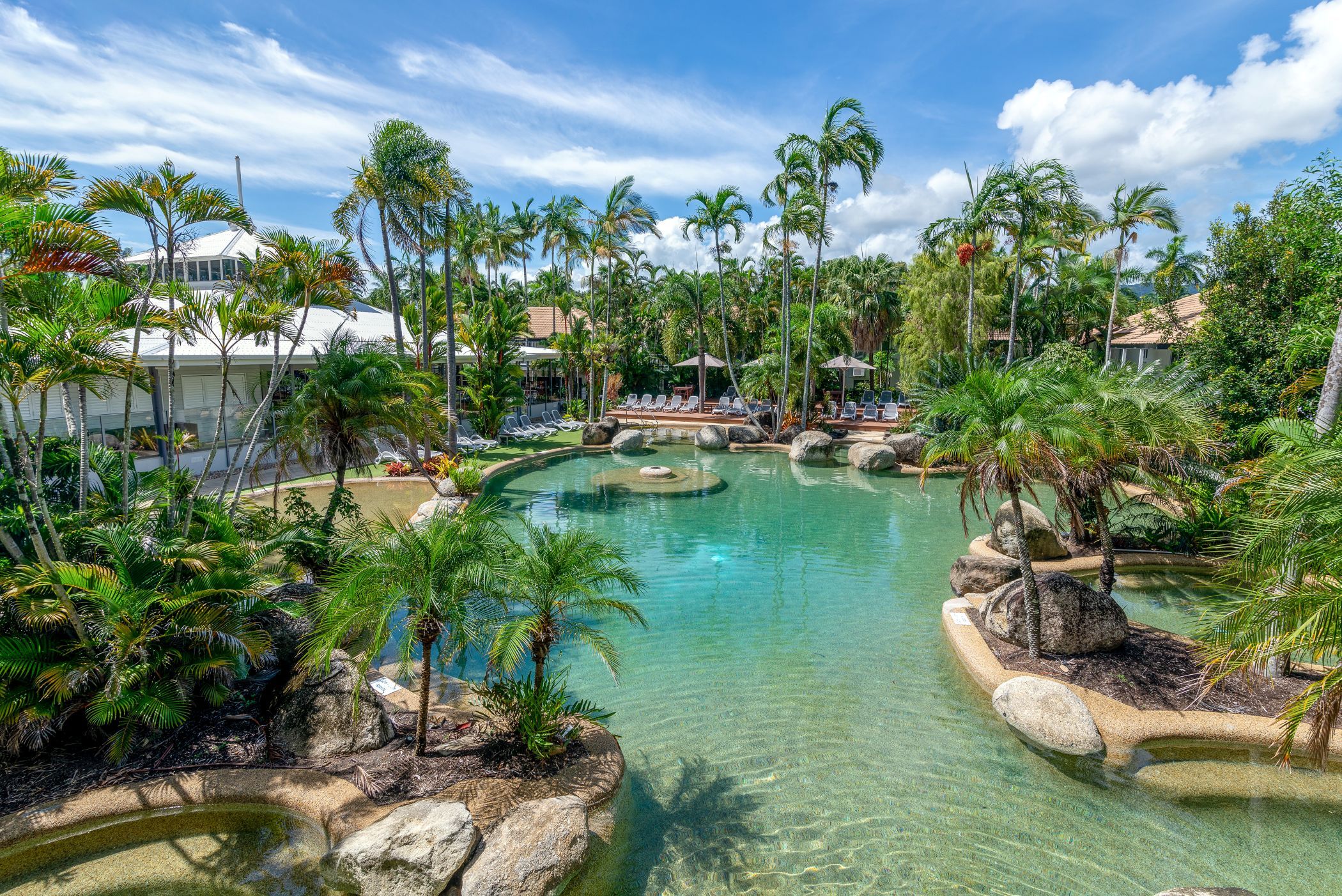 A resort with a clear pool surrounded by palm trees and lounge chairs.