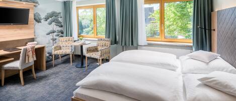 Una habitación de hotel con una cama, un escritorio con una silla, una pequeña mesa con sillas, un televisor montado en la pared y ventanas amplias con cortinas.