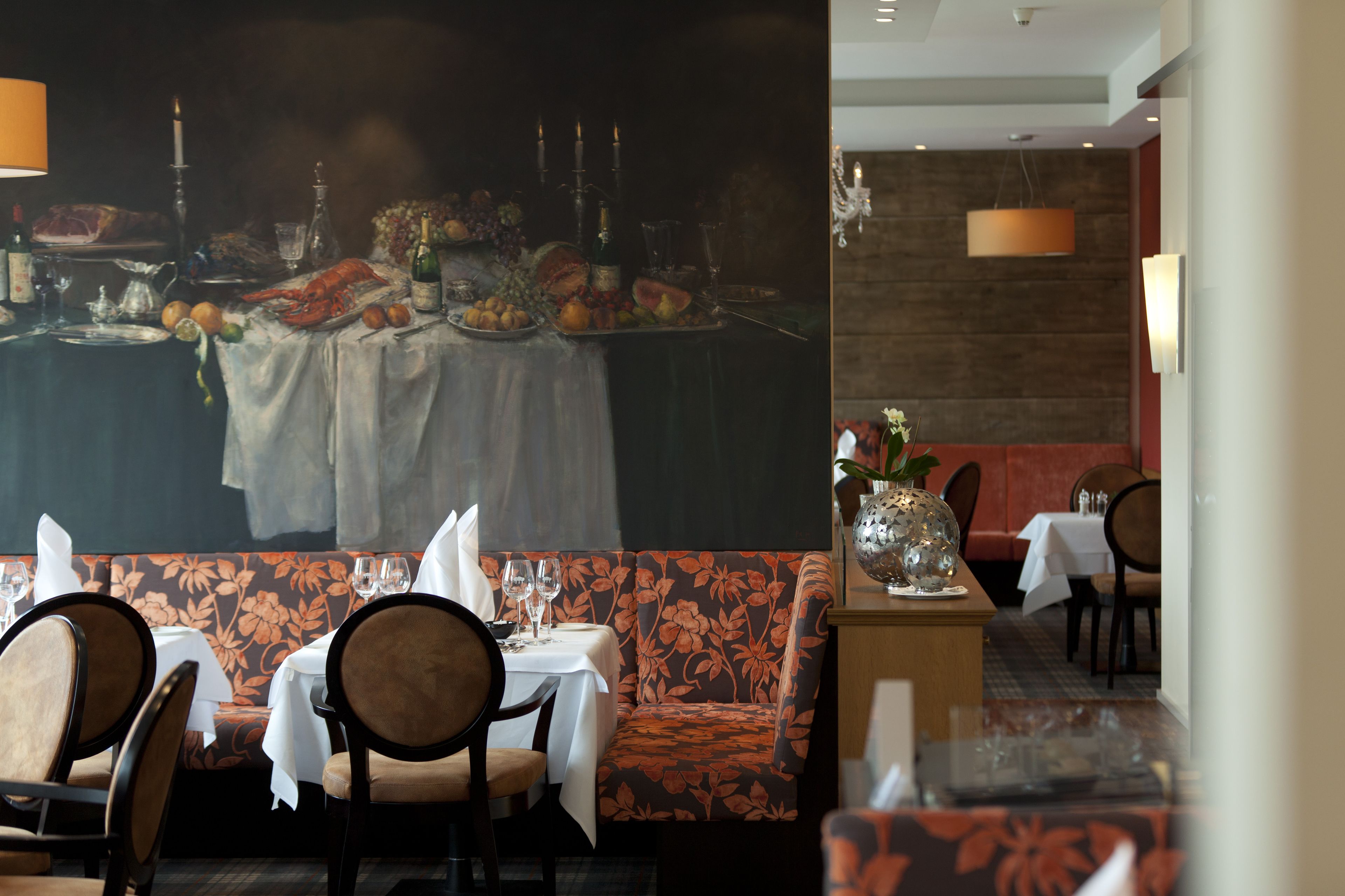 En restaurant med et spiseområde, hvor borde er dækket til to personer, et stort blomstermaleri på væggen og en mønstret bænk.