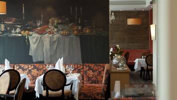 Un restaurante con un comedor que presenta mesas dispuestas para dos personas, un mural floral grande en la pared y un banco con estampado.
