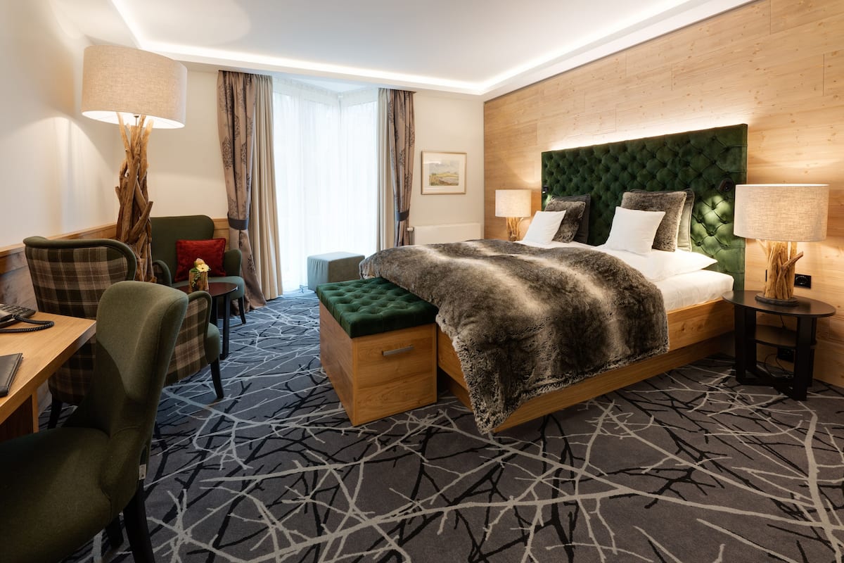 A hotel room with a large bed, a desk, a chair, a small table, and a lamp.