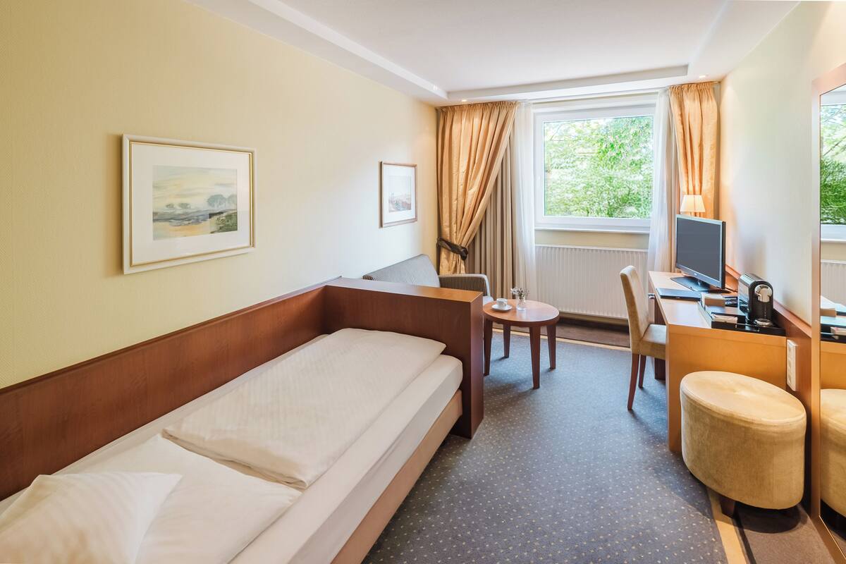 A hotel room with a bed, a desk with a computer, a chair, and a window with curtains.