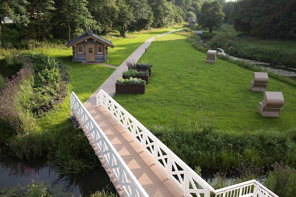 A wooden bridge leading to a rustic cabin with a small porch, surrounded by lush greenery and a calm water body. - Romantik Hotel Waldschlösschen (Schleswig)