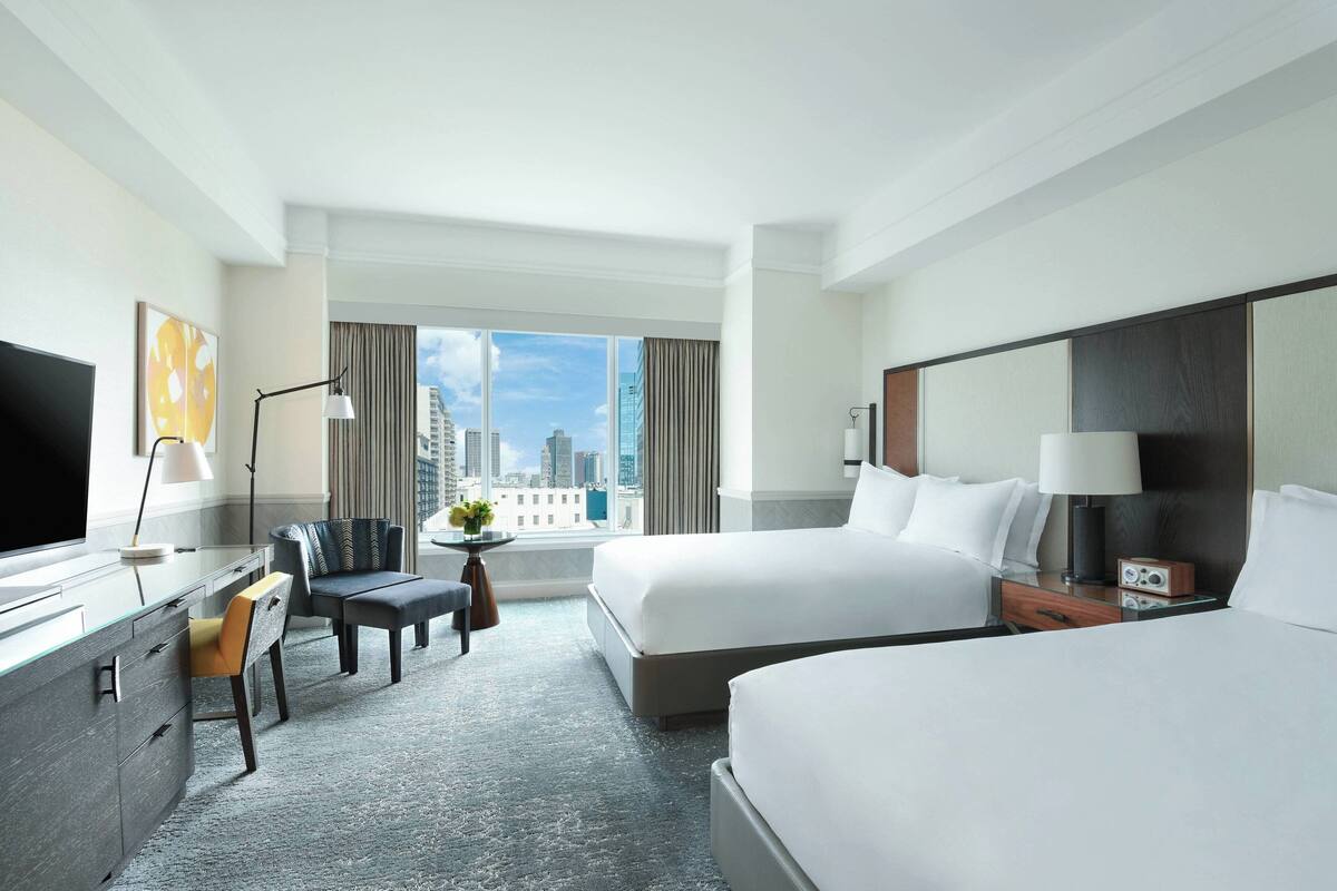 Une chambre d’hôtel avec deux lits, un bureau avec une chaise, une télévision et une grande fenêtre donnant sur la ville.