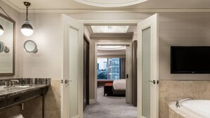 A modern hotel bathroom with a marble vanity, a large bathtub, and a city view through an open door.