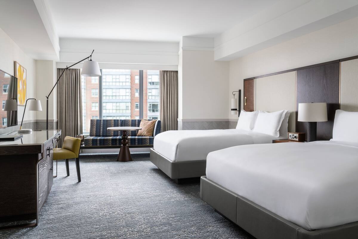 Une chambre d’hôtel avec deux lits, un bureau, une chaise, une lampe et une vue sur les immeubles par la fenêtre.
