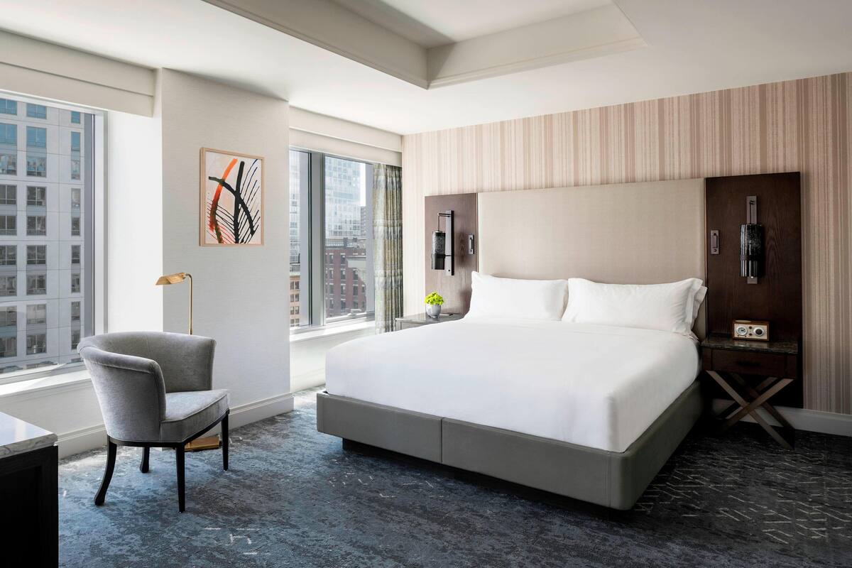 Une chambre d’hôtel moderne dotée d’un grand lit, d’une chaise, d’un bureau et offrant une vue sur la ville grâce à de grandes fenêtres.