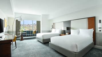 A hotel room with two beds, a desk, a chair, a lamp, and a view of the city.