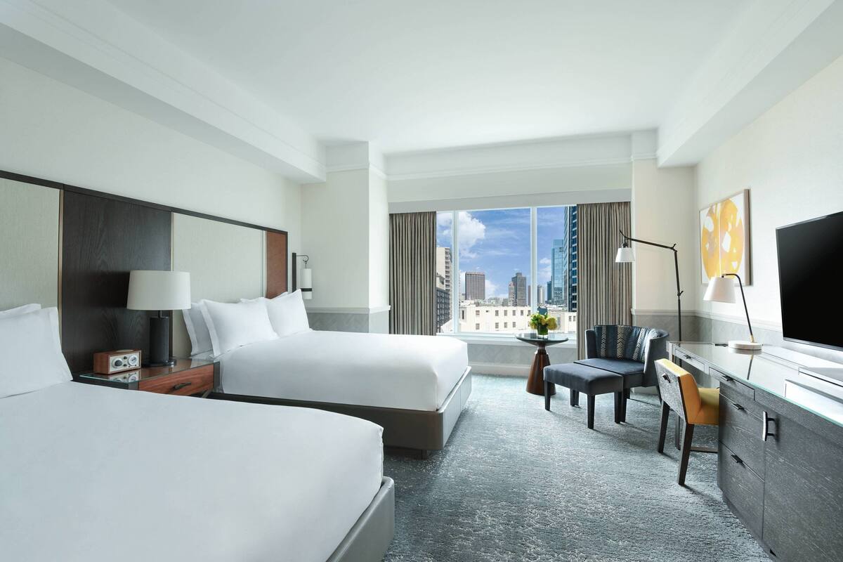 Une chambre d’hôtel avec deux lits, un bureau, une chaise, une télévision et une grande fenêtre donnant sur la ville.