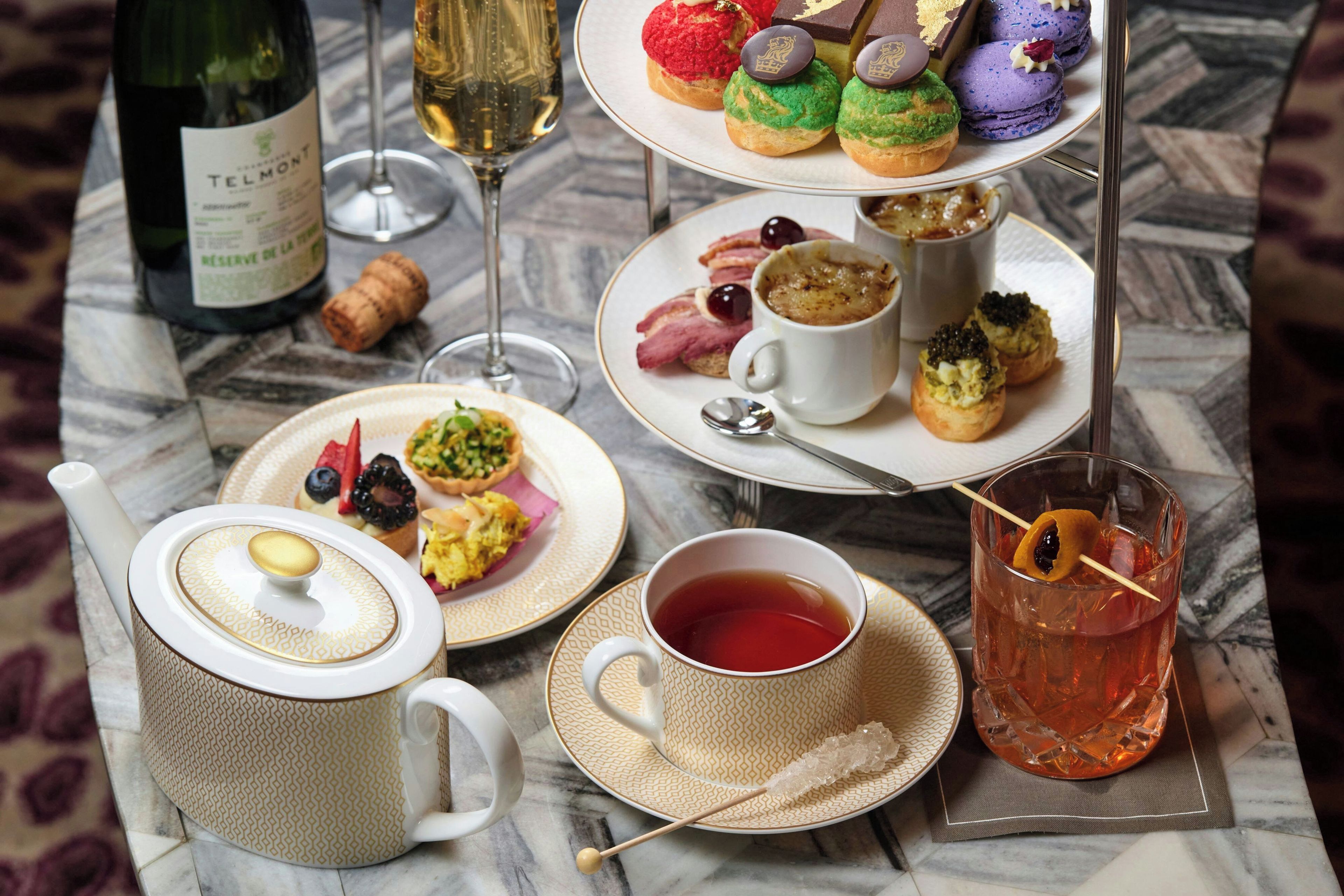 Une table dressée avec une bouteille de vin, des verres, une théière, des tasses, un plateau de pâtisseries et un verre de thé.