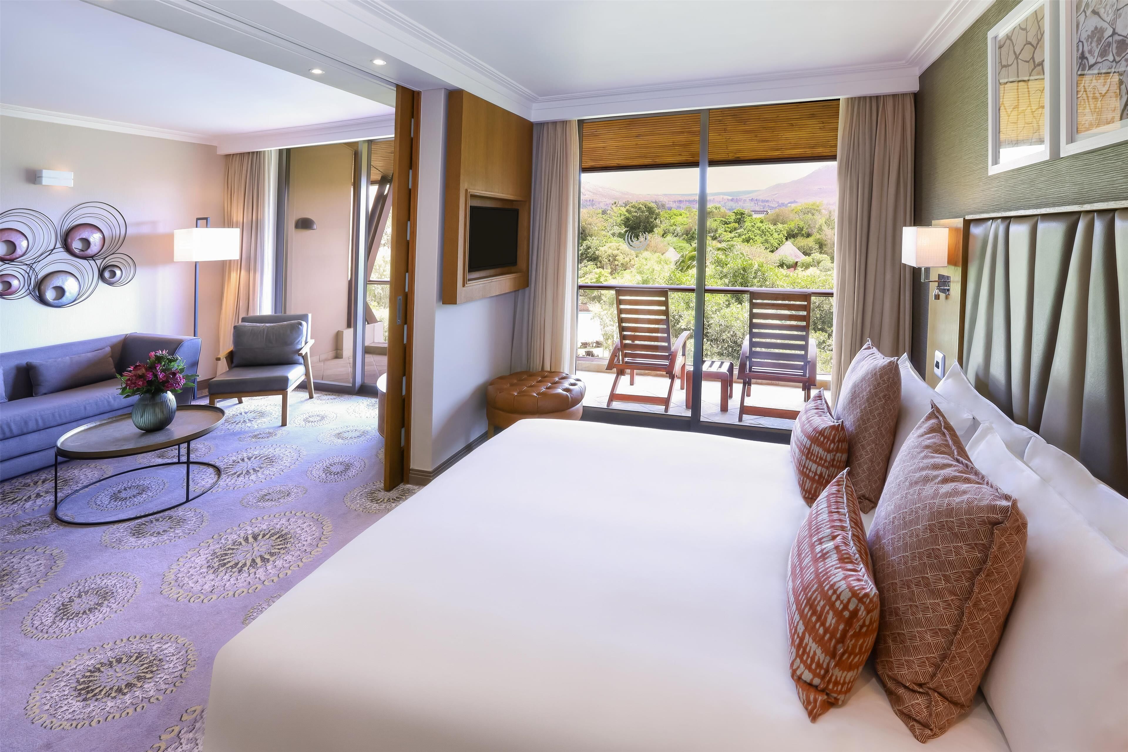 A hotel room with a large bed, a sofa, a coffee table, and a balcony with a view of the mountains.