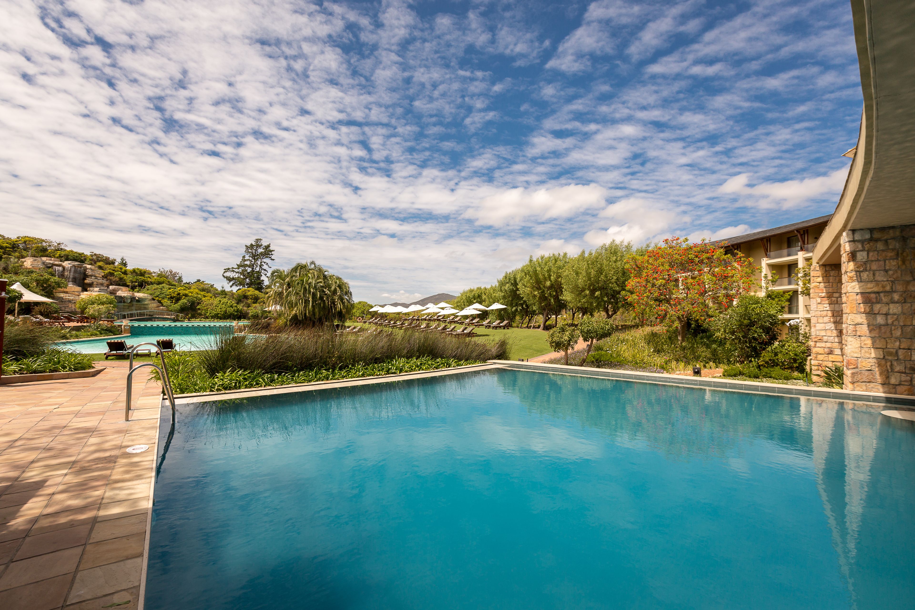 A swimming pool with a clear sky, surrounded by greenery and a stone building.