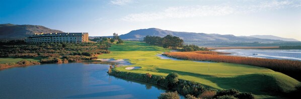 A golf course with a large building on the left, surrounded by mountains and a body of water. - Arabella Hotel Golf & Spa (Kleinmond)