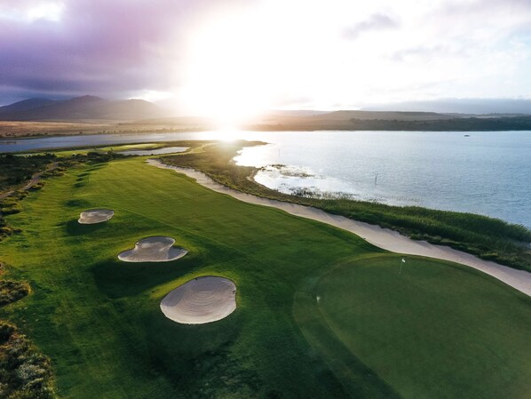Campo de golf con búnkeres, una masa de agua y un atardecer.
