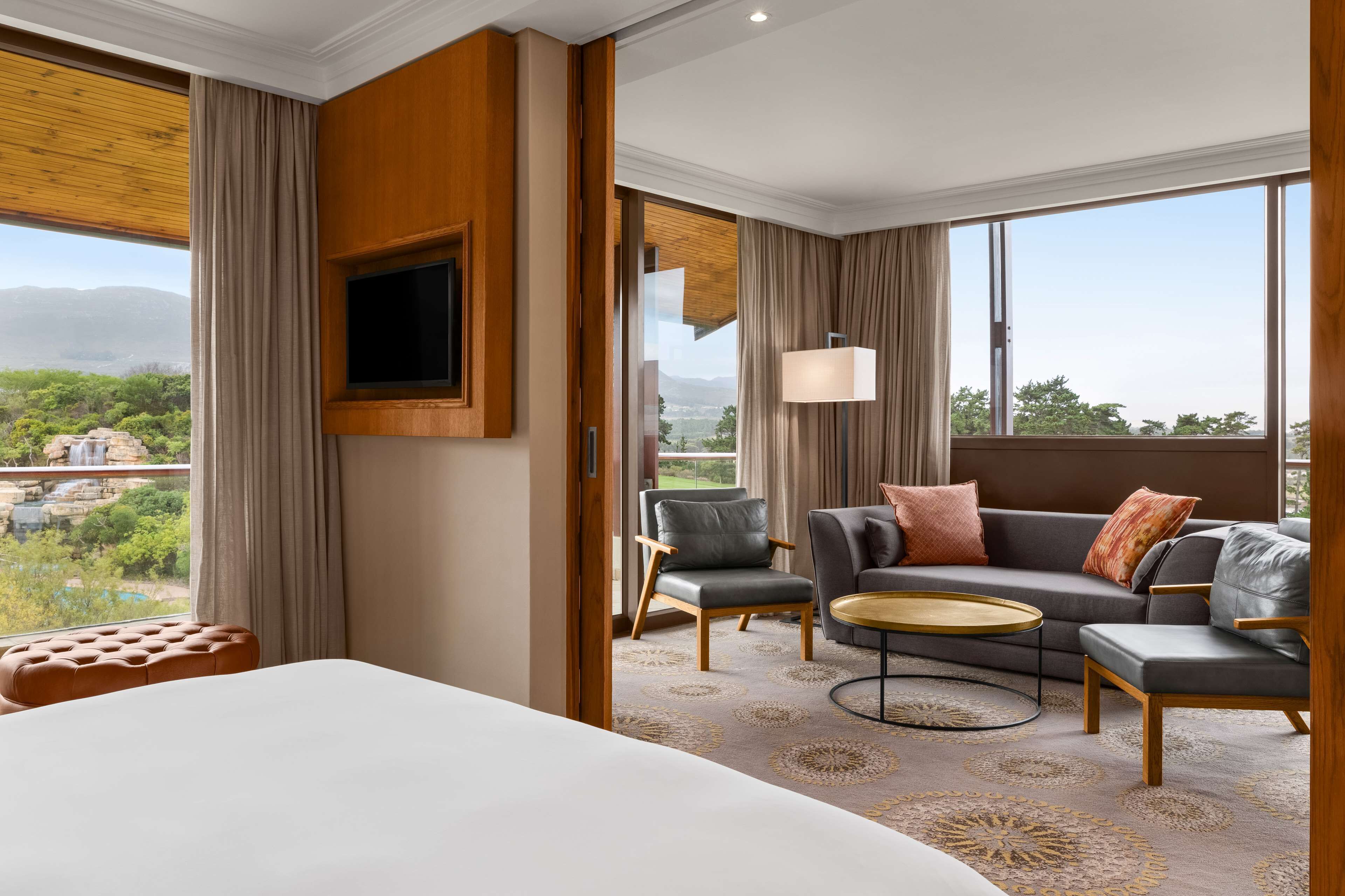 A hotel room with a large bed, a sofa, two armchairs, a small round table, and a TV mounted on the wall.