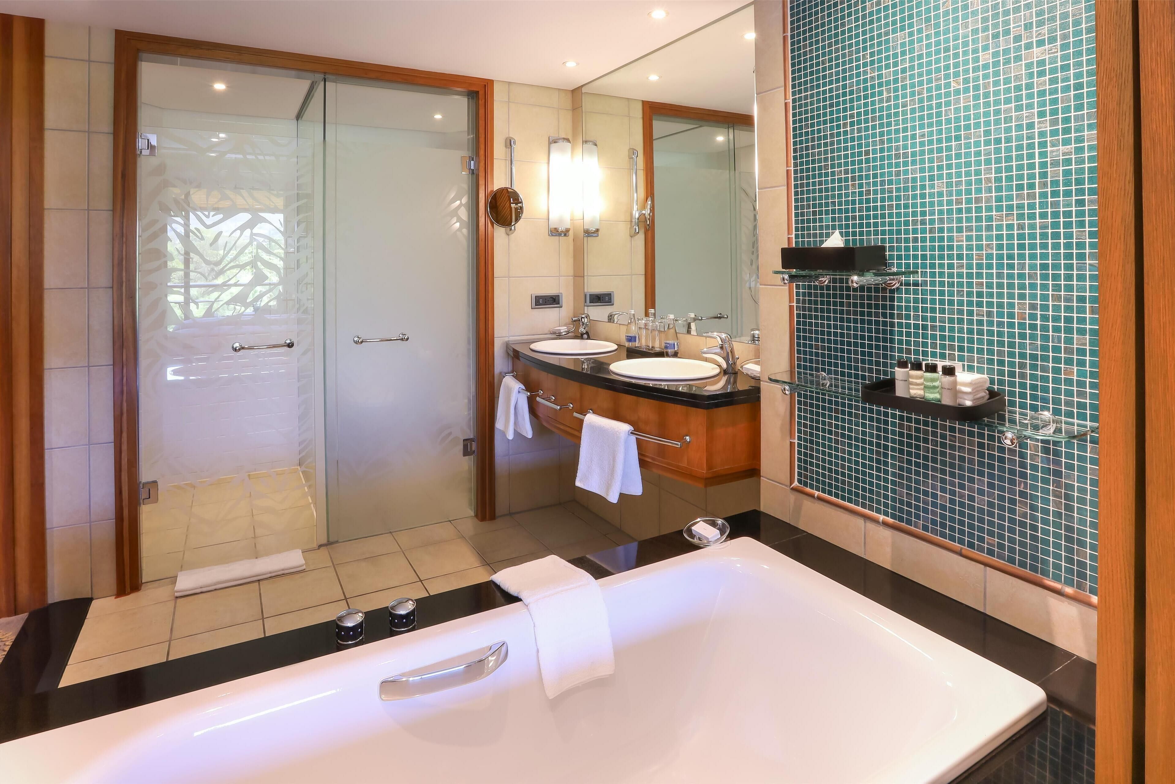 A modern bathroom with a large bathtub, two sinks, a glass shower enclosure, and a tiled wall.