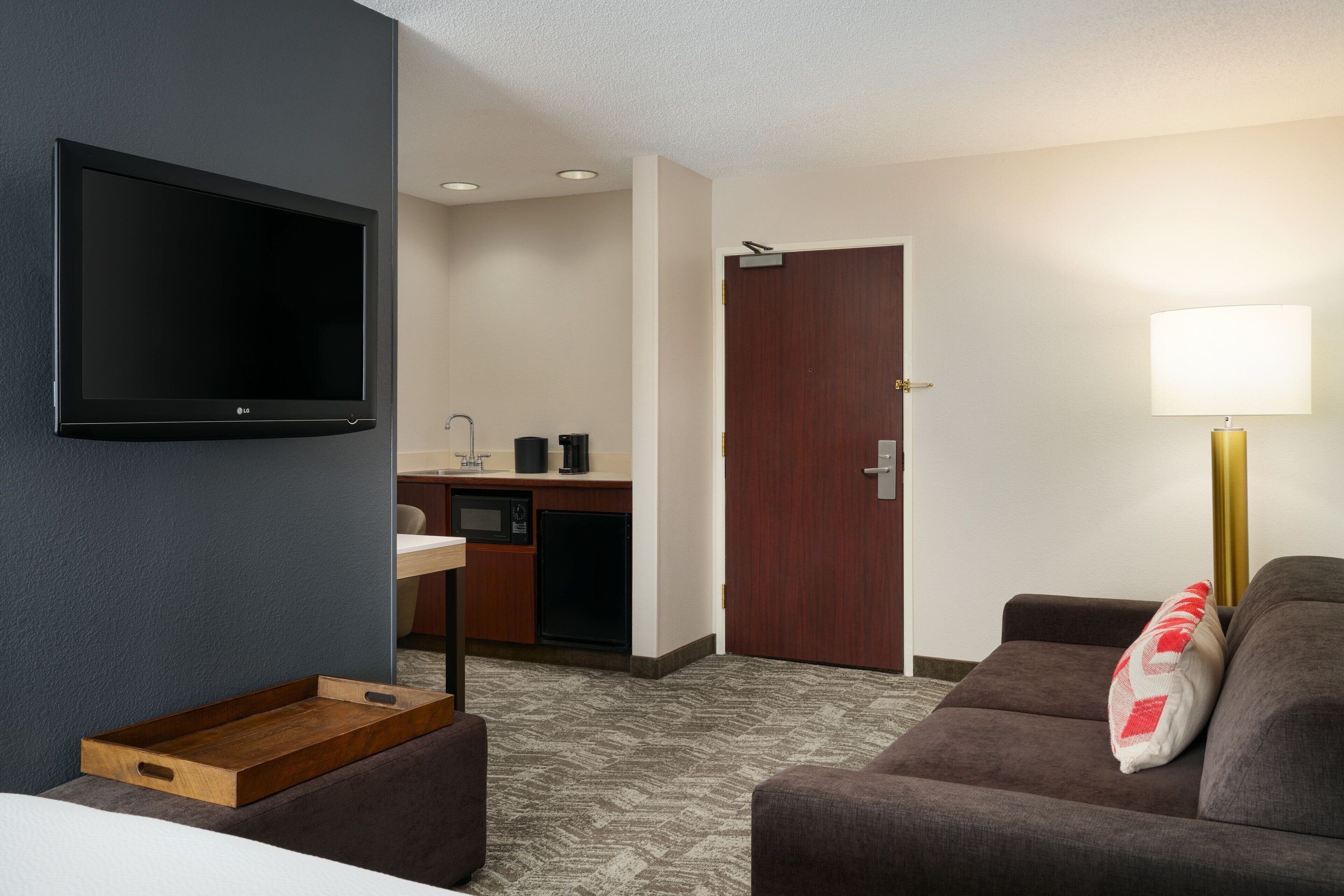 A hotel room with a flat-screen TV, a sofa, a wooden tray, a kitchenette, and a door.