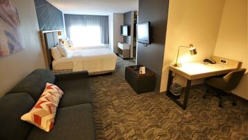 A hotel room with a bed, a desk with a lamp, a TV, a couch, and a trash bin.