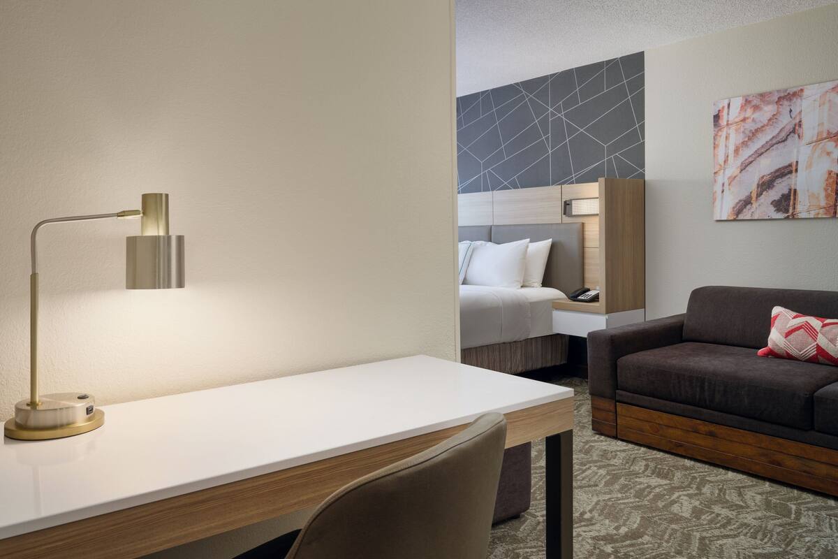 Habitación de hotel con escritorio, lámpara, cama, sofá y una pared decorada con arte abstracto.
