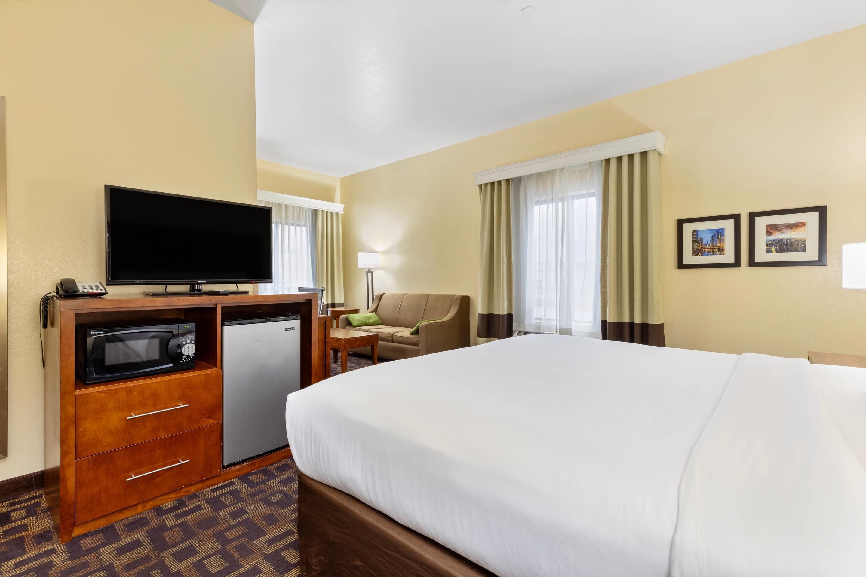 A hotel room with a bed, a TV, a microwave, a mini-fridge, a desk with a lamp, a chair, and two framed pictures on the wall.