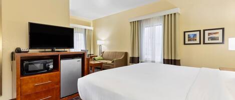 A hotel room with a bed, a TV, a microwave, a mini-fridge, a desk with a lamp, a chair, and two framed pictures on the wall.