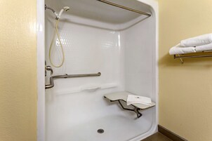 Un box de ducha blanco con ducha de mano, una barra de apoyo y un estante pequeño con una toalla.