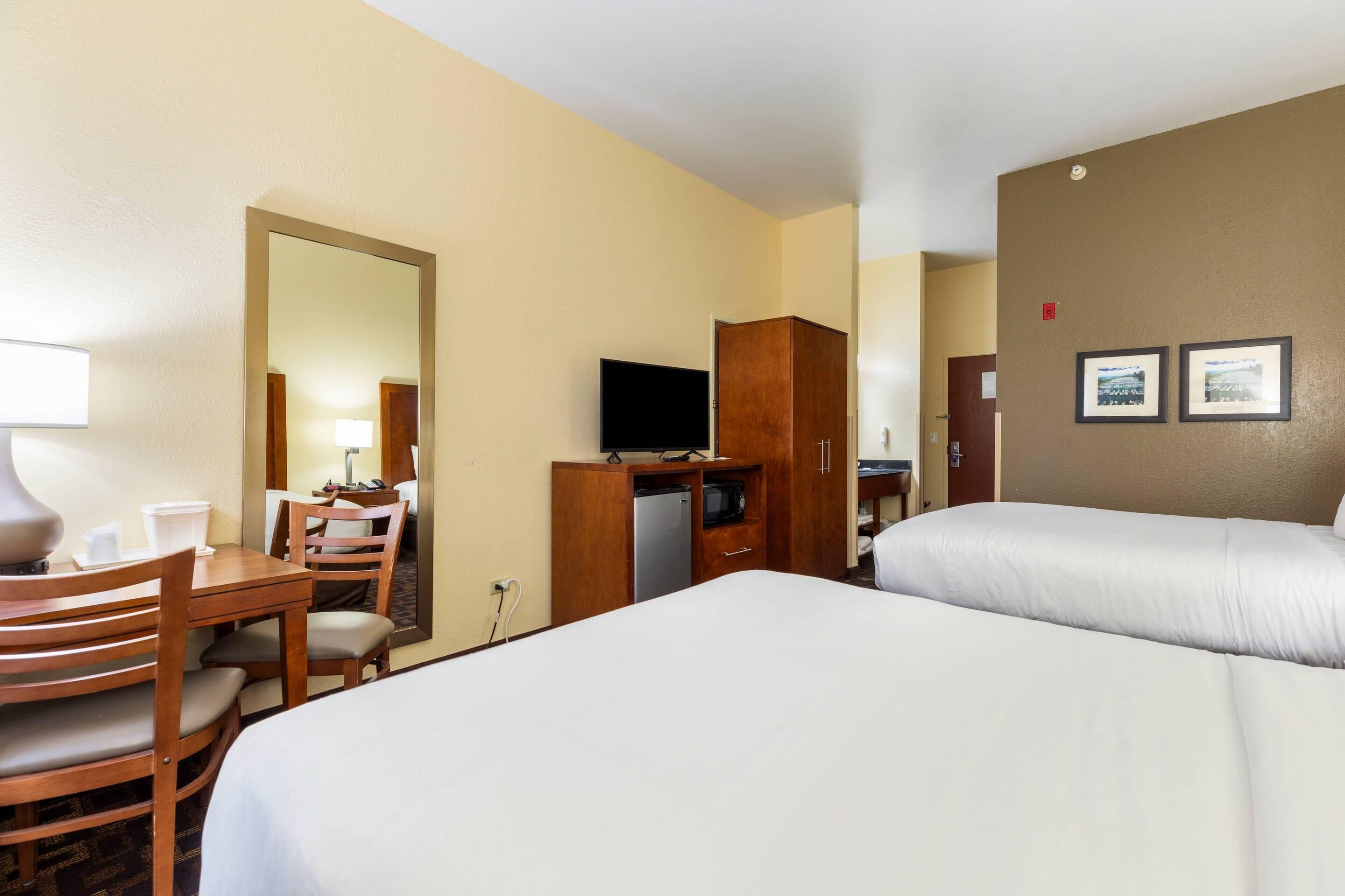 Habitación de hotel con dos camas, un escritorio con silla, televisor, minibar y espejo.