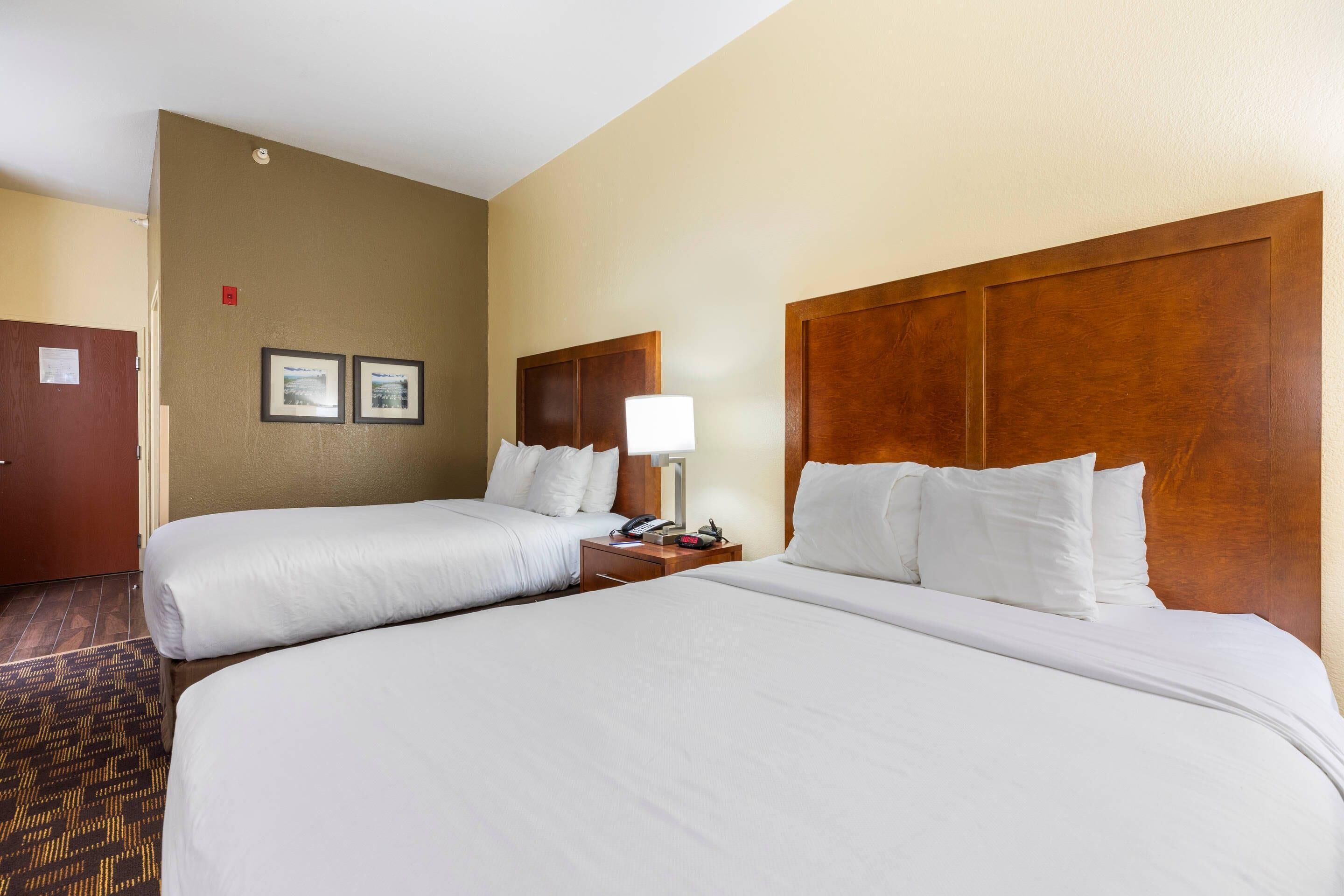 Quarto de hotel com duas camas, cabeceira de madeira, mesa de cabeceira com abajur e piso coberto por carpete.