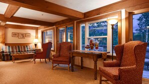 A cozy living room with wooden beams, a sofa, armchairs, a table with decorative items, and large windows offering a view of the outdoors.