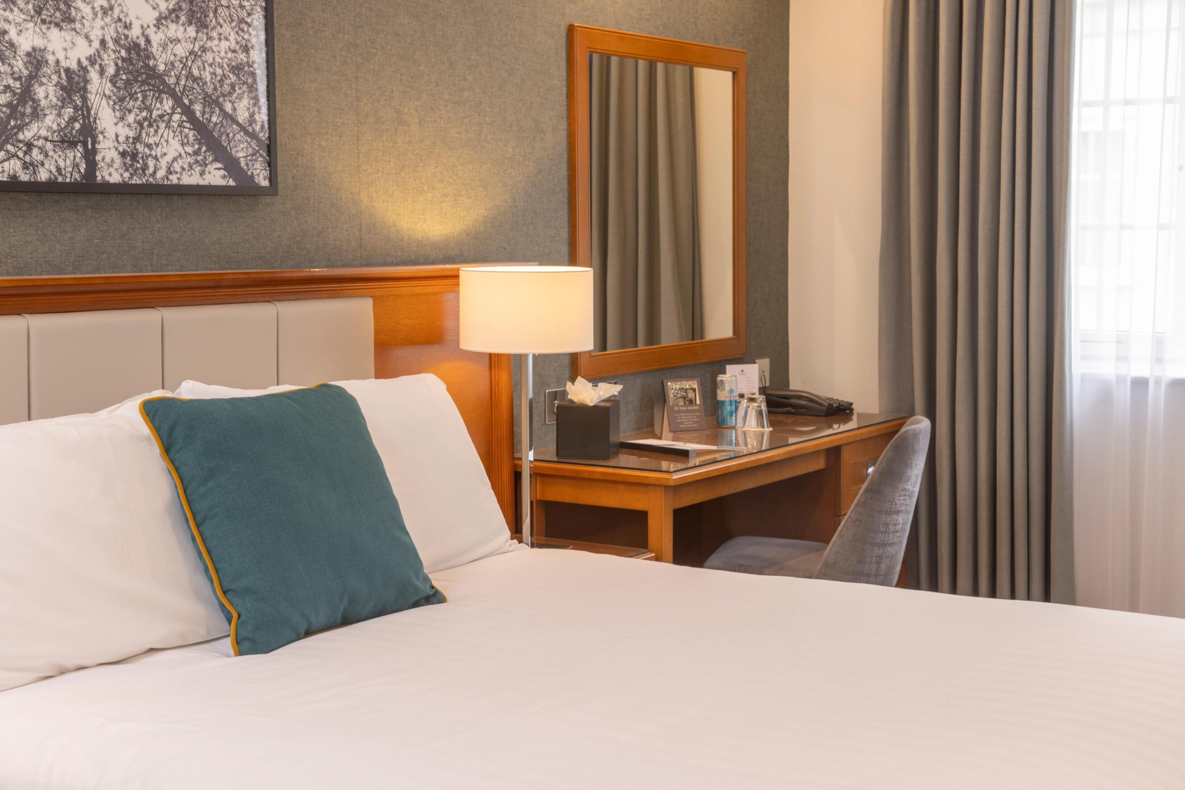 A hotel room with a bed, a desk, a chair, a lamp, a mirror, and a window with curtains.