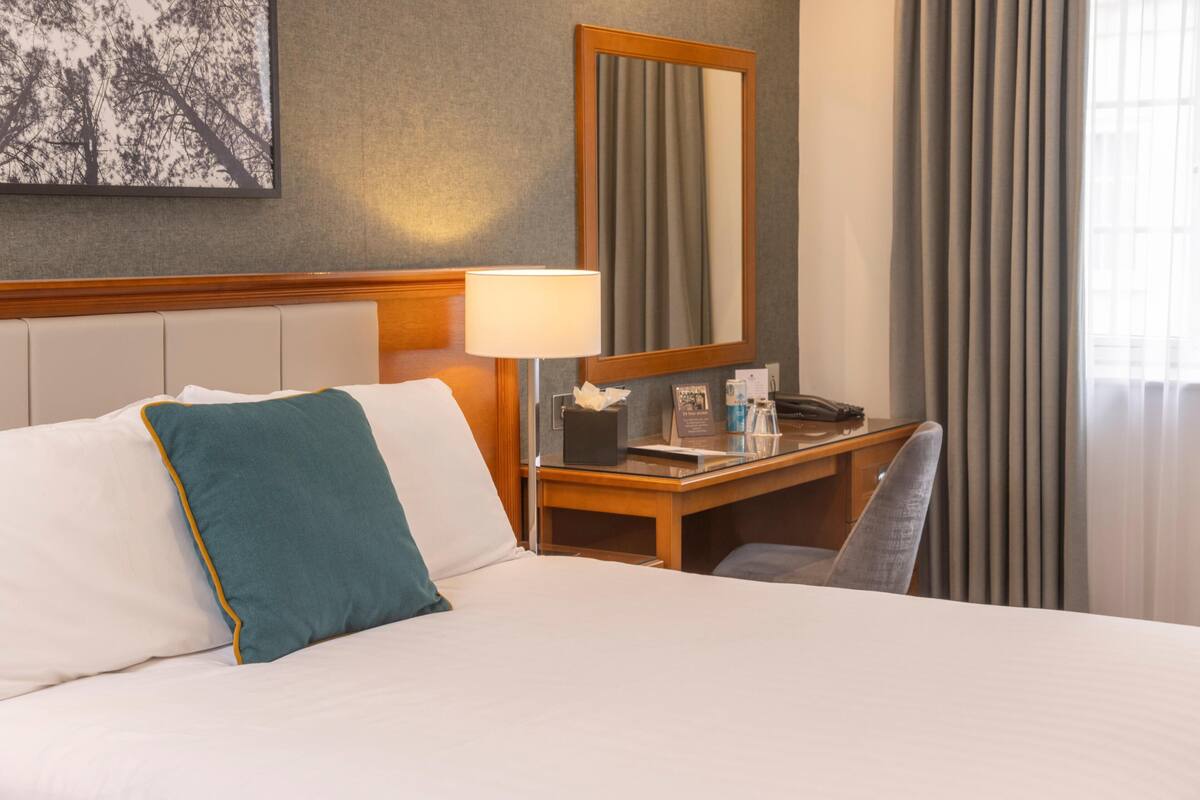 Una habitación de hotel con cama, escritorio, silla, lámpara, espejo y ventana con cortinas.